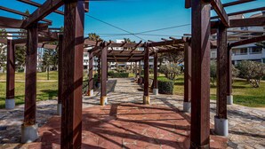 Une structure de pergola en bois, dotée d’un sol en pierre, entourée de verdure et avec des bâtiments en arrière-plan.