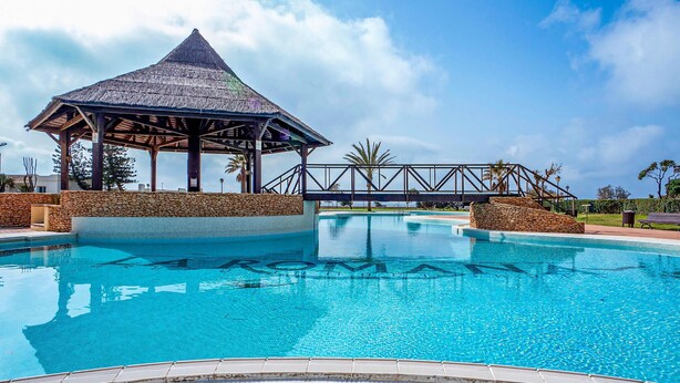 Une piscine agrémentée d’un kiosque, de murs en pierre et surplombée d’un ciel bleu clair.