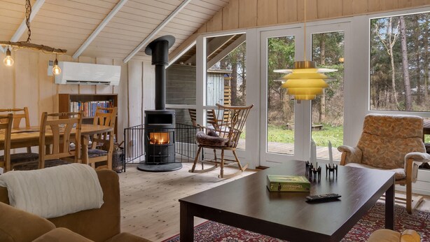 A cozy living room with a wood-burning stove, wooden furniture, and large windows offering a view of the outdoors. - Stunning home in Gedser with sauna (Gedser)