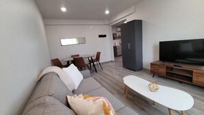 Un salon moderne avec un canapé gris, une table basse blanche et un téléviseur à écran plat posé sur un meuble en bois. On aperçoit un coin repas avec une table et des chaises, ainsi qu’une cuisine au fond.