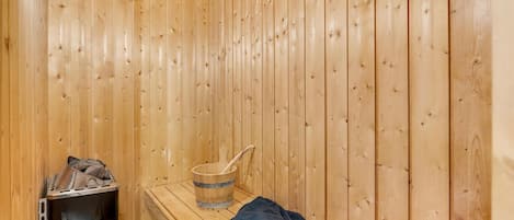 A sauna room with wooden walls, a bench, a bucket, and a sauna heater.