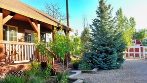 Une cabane en bois dotée d’un porche, entourée d’une allée en gravier et d’un petit jardin où poussent diverses plantes et un arbre à feuilles persistantes.