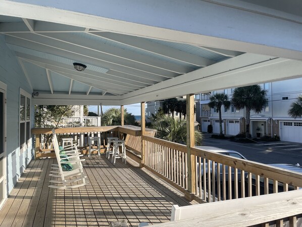 A covered wooden deck with a railing, a wooden table, and several white chairs.