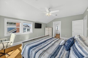 A bedroom with a bed, a desk, a chair, a ceiling fan, and a window with a view of a house.