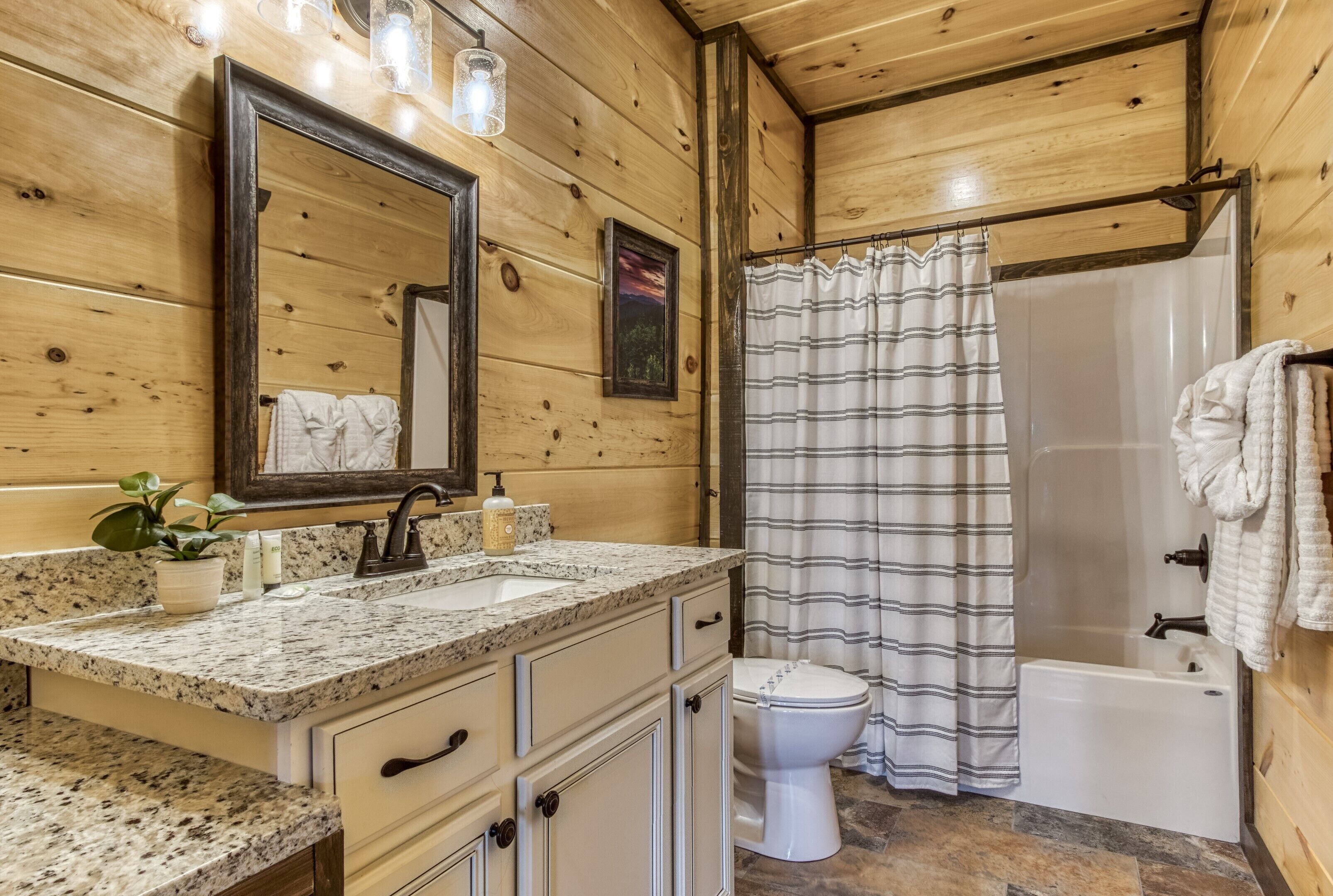 A wooden cabin bathroom with a granite countertop sink, a white toilet, a shower with a striped curtain, and a mirror with lights above it.