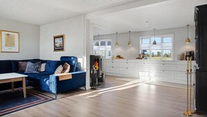 Una sala de estar moderna con un sofá azul, una chimenea y una cocina con armarios blancos y lámparas colgantes.