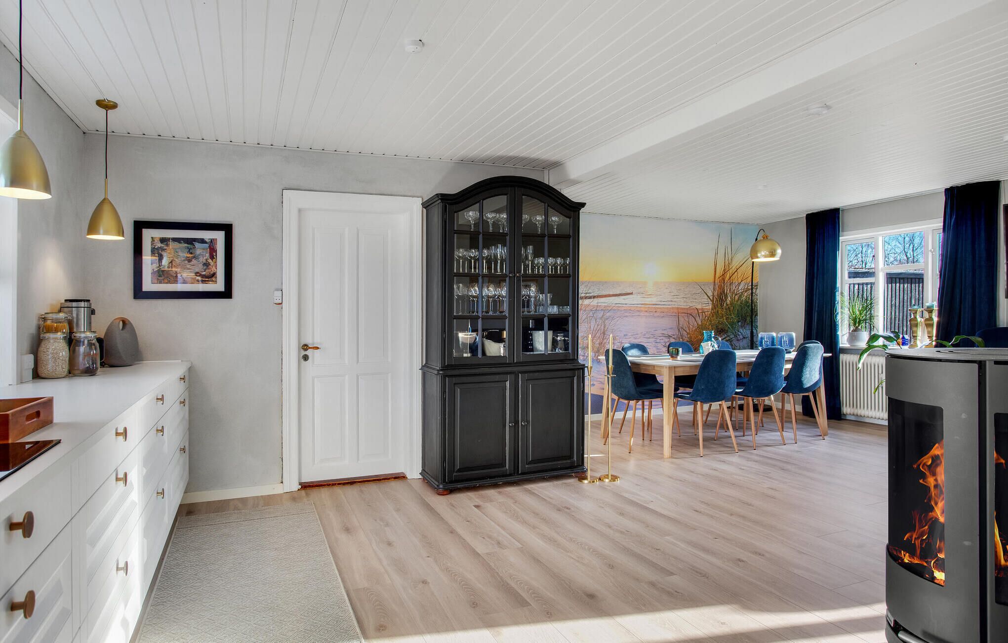 A modern kitchen with white cabinets, a black cabinet with glassware, a dining area with blue chairs, and a large window with a view of the sea.