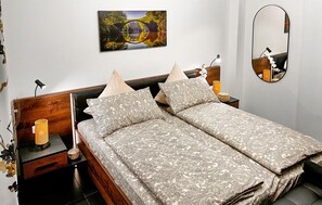 A double bed with patterned bedding, a nightstand with a lamp, a wall-mounted mirror, and a painting above the bed.