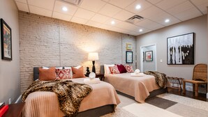 A hotel room with two beds, a brick wall, a large painting, and a chair. - YOUR URBAN OASIS - Paristown, Downtown, Highlands, NULU - brand new!! (Louisville)