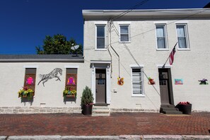A two-story white building with a brick sidewalk, a small entrance with steps, and a flag on the right side.