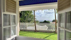 A view from a window with white shutters, looking out to a white picket fence, green lawn, and palm trees. - Welcome to the heart of my island (Schoelcher)