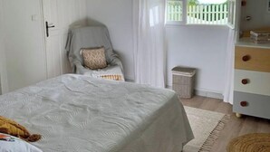 A bedroom with a bed, a bedside table, a dresser, a window with curtains, a hanging light fixture, and a door. - Welcome to the heart of my island (Schoelcher)