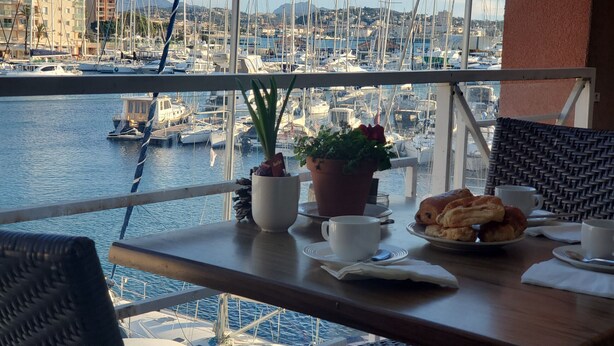 A balcony with a table set for breakfast, overlooking a marina with boats and buildings in the background.
