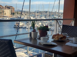 Un balcon avec une table préparée pour le petit-déjeuner, donnant sur une marina où l’on aperçoit des bateaux et des bâtiments en arrière-plan.