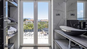 Ein modernes Badezimmer mit großem Fenster, einem schwarzen Marmorwaschtisch und Blick auf einen Balkon mit Stadtansicht.