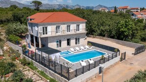 Ein zweistöckiges Gebäude mit Swimmingpool, umgeben von Grünflächen und Bergen im Hintergrund.