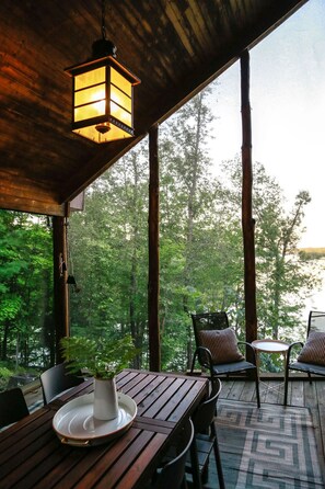 A wooden deck with a table and chairs, a hanging lantern, and a view of a forested area. - Stylish and Cozy Cabin on the Red Cedar Chain overlooking quiet bay (Birchwood)