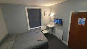 Habitación pequeña con cama individual, ventana, mesita de noche, televisor y puerta con un cartel de seguridad contra incendios.