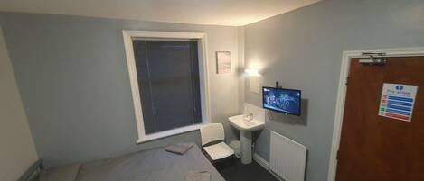 A small, single-bed room with a window, a bedside table, a TV, and a door with a fire safety sign.