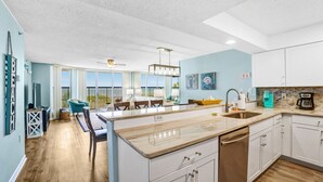 Une cuisine moderne avec des meubles blancs, un grand îlot central et un coin repas agrémenté d’une chaise bleue.