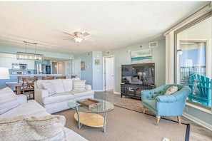A spacious living room with a sofa, armchairs, and a glass coffee table. A kitchen is visible in the background, and there is a large window with a view.