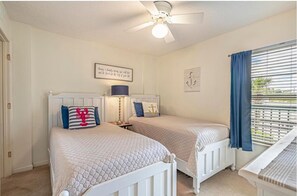 A bedroom with two beds, a ceiling fan, a window with curtains, and a wall with a framed quote.