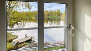 Ein Fenster mit Blick auf einen See, eine Holzsteg und eine Wiese mit Bäumen.