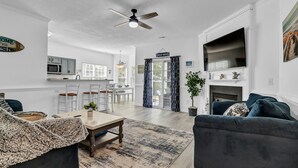 A spacious living room with a dark gray sofa, a wooden coffee table, and a patterned rug. A ceiling fan is visible, and there is a kitchen area in the background with a microwave and cabinets. - Beach House Fun Time (Wilmington)