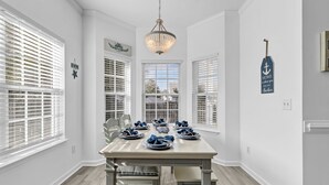 A dining area with a white table set for six, large windows with blinds, a chandelier, and nautical decor. - Beach House Fun Time (Wilmington)