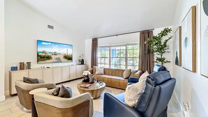 A modern living room with a large flat-screen TV, comfortable seating, a coffee table, and a view of the outdoors through large windows. - Palma Sola Getaway | Private Pool + Games (Bradenton)