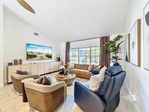 A modern living room with a large flat-screen TV, comfortable seating, a coffee table, and a view of the outdoors through large windows.