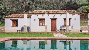A two-story house with a tiled roof, a swimming pool in front, and a small patio with outdoor furniture.