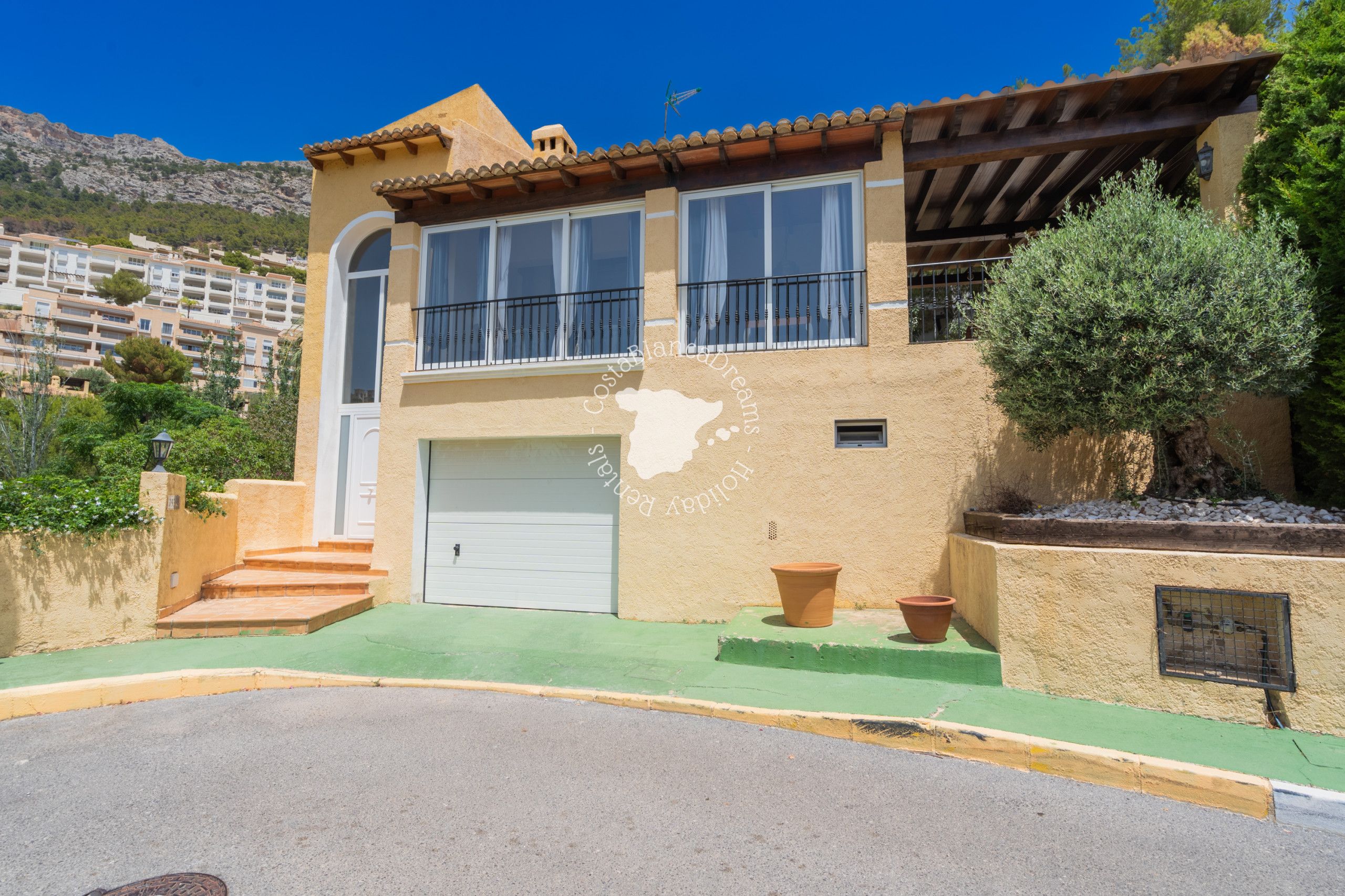 Una casa de dos pisos con garaje, balcón y montañas al fondo.