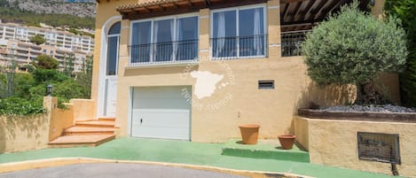 Una casa de dos pisos con garaje, balcón y montañas al fondo.