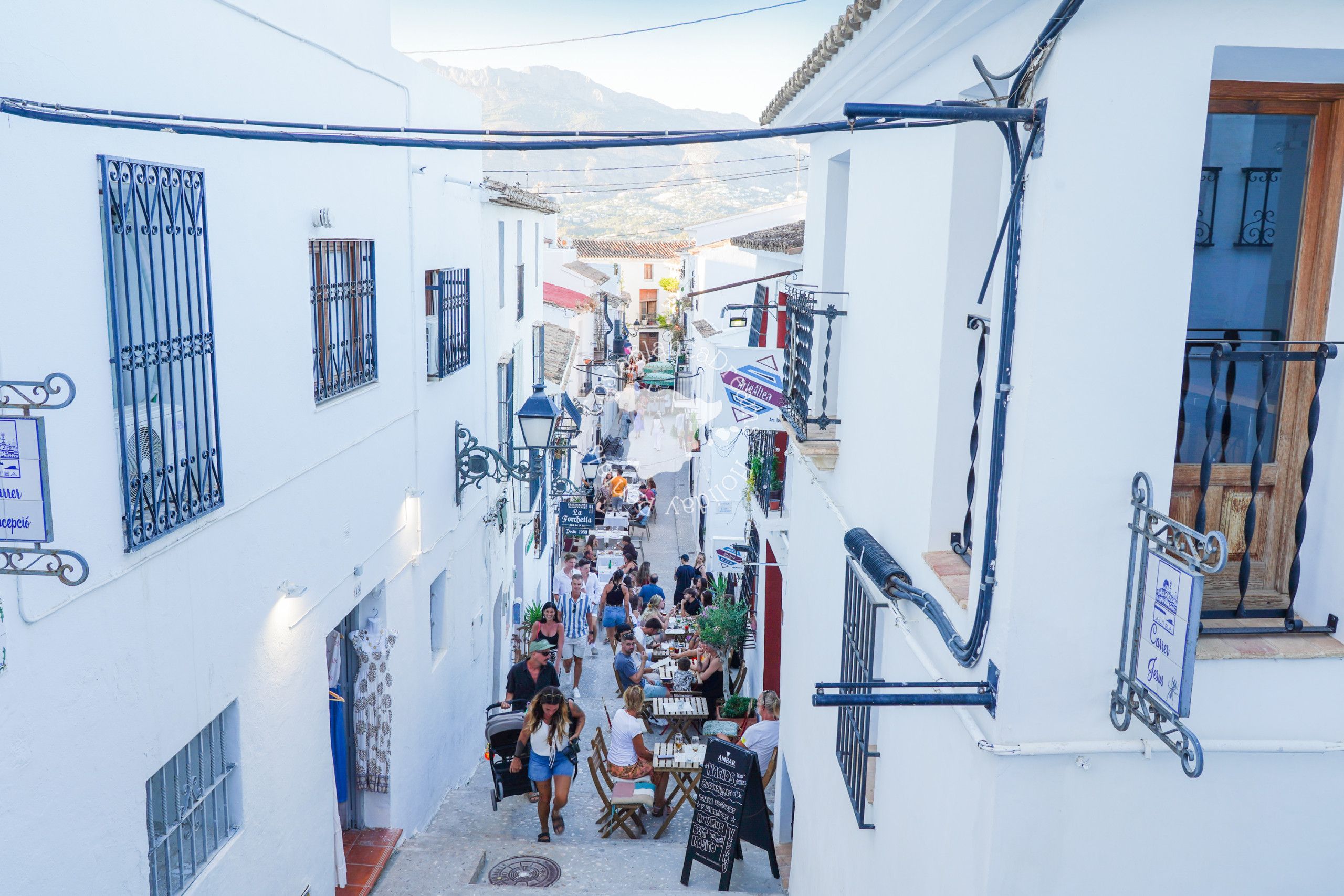 A narrow street with white buildings, outdoor seating, and a signboard.
