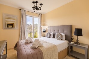 A bedroom with a large bed, a bedside table with a lamp, a mirror, and a window with curtains.