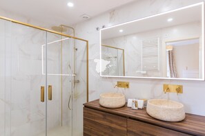 A modern bathroom with a glass-enclosed shower, marble walls, and two round sinks with gold fixtures.