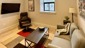A modern living room with a flat-screen TV mounted on the wall, a leather armchair, a sofa, a wooden coffee table, and a window with a view of buildings. - Two bedroom suite - great for visiting family, weddings, or Stratford Festival (St. Marys)
