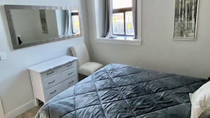 A bedroom with a large bed, a dresser, a chair, a window with curtains, and a mirror on the wall. - Two bedroom suite - great for visiting family, weddings, or Stratford Festival (St. Marys)