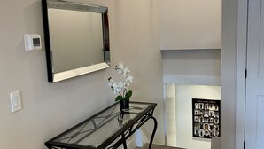 A hallway with a glass-top console table, a wall-mounted mirror, a small potted orchid, a wall-mounted light fixture, and a framed picture on the wall. - Two bedroom suite - great for visiting family, weddings, or Stratford Festival (St. Marys)