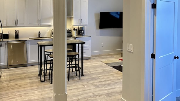 A modern kitchen with white cabinets, a dining area with a table and chairs, and a large TV mounted on the wall. - Two bedroom suite - great for visiting family, weddings, or Stratford Festival (St. Marys)