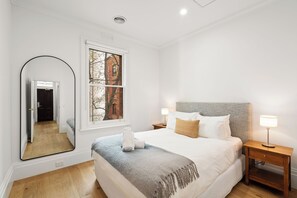 A bedroom with a large bed, a nightstand, a mirror, and a window with a view of a building. - Gordon House Apartments (Melbourne)