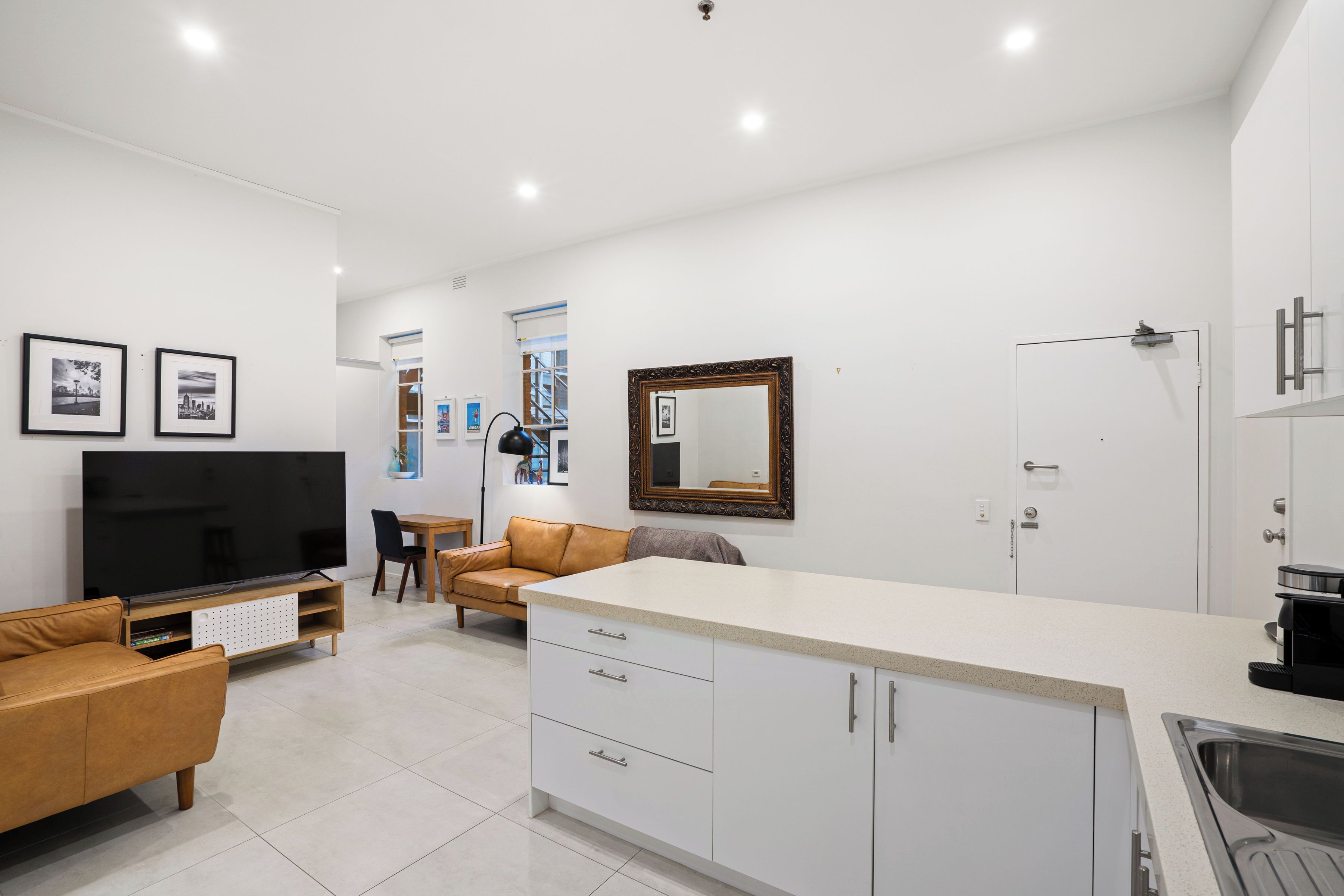 Una cocina moderna con armarios blancos, una isla grande y un horno empotrado. Hay un comedor con mesa y sillas, y una sala de estar con sofá y televisor.