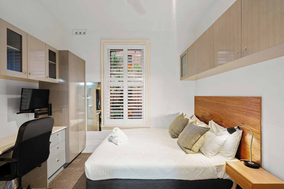 A bedroom with a bed, bedside table, TV, desk, and wardrobe.