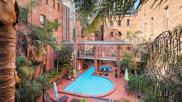 Uma piscina no terraço com cobertura de vidro, cercada por palmeiras e edifícios de tijolo.