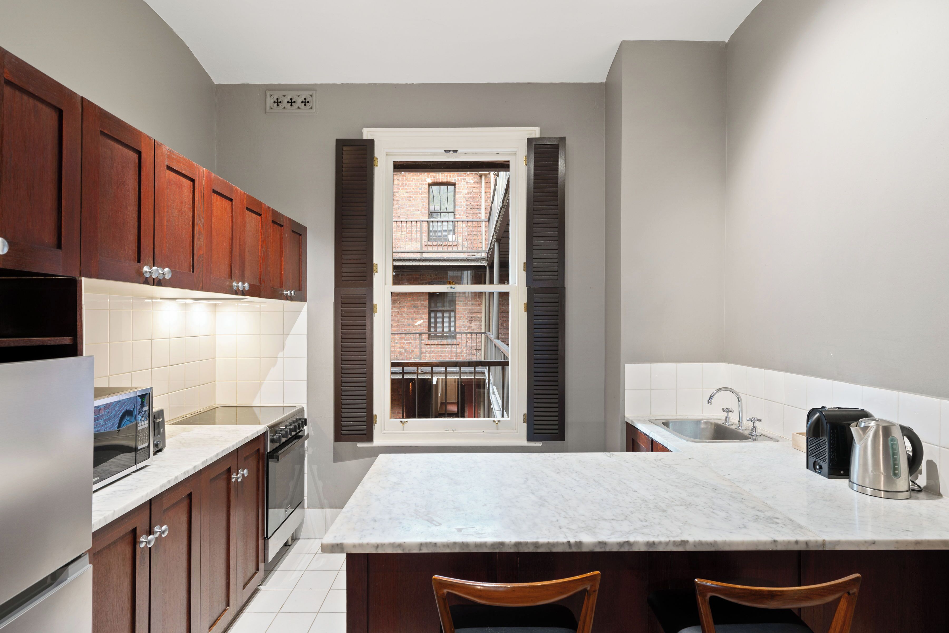 A kitchen with wooden cabinets, a marble countertop, a window with shutters, and a view of a brick building.