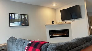 Una sala de estar moderna con un sofá gris, una chimenea, un televisor montado y un cuadro con un paisaje nevado.