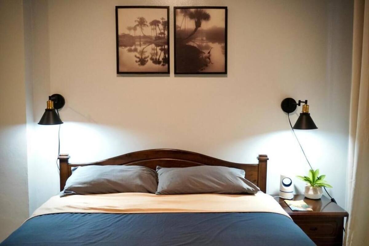 Una cama bien hecha con cabecera de madera, dos lámparas fijadas a la pared y dos cuadros enmarcados.