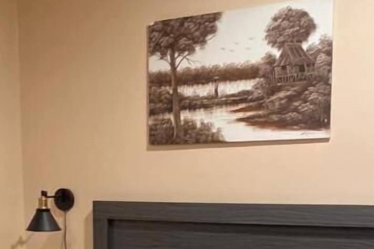 Un dormitorio con cama, almohadas, cabecera, lámpara de pared y un cuadro.