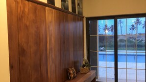 A wooden bench with a cushion and a small plant, a large window with a view of palm trees and a pool, and a wall with three framed pictures.